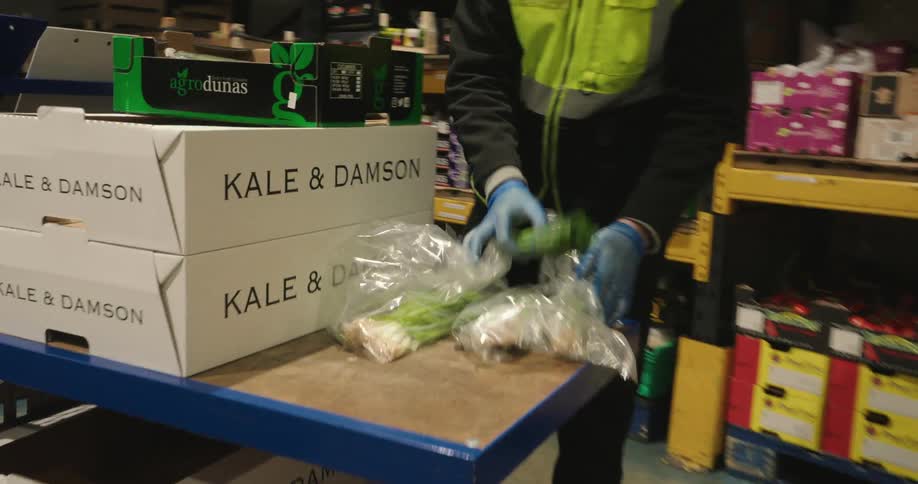 A man in a safety vest and wearing a hat loading a cart with vegetables in a distribution warehouse