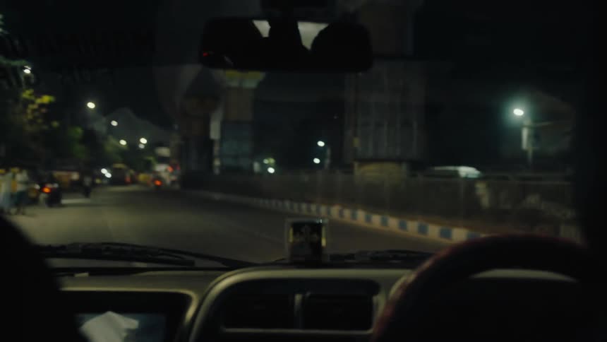 A nighttime urban street scene viewed from inside a moving vehicle featuring pedestrians parked vehicles and an elevated metro structure under construction