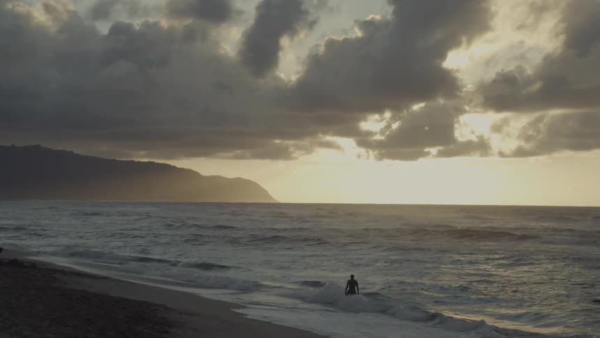 A man stands in ocean waves on a beach at sunset