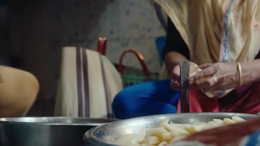 Woman slices food while sitting on a low seat in front of a large metal bowl