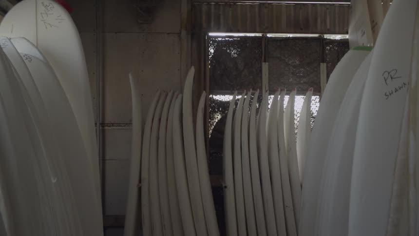Unfinished surfboards line the interior of a warehouse during the day