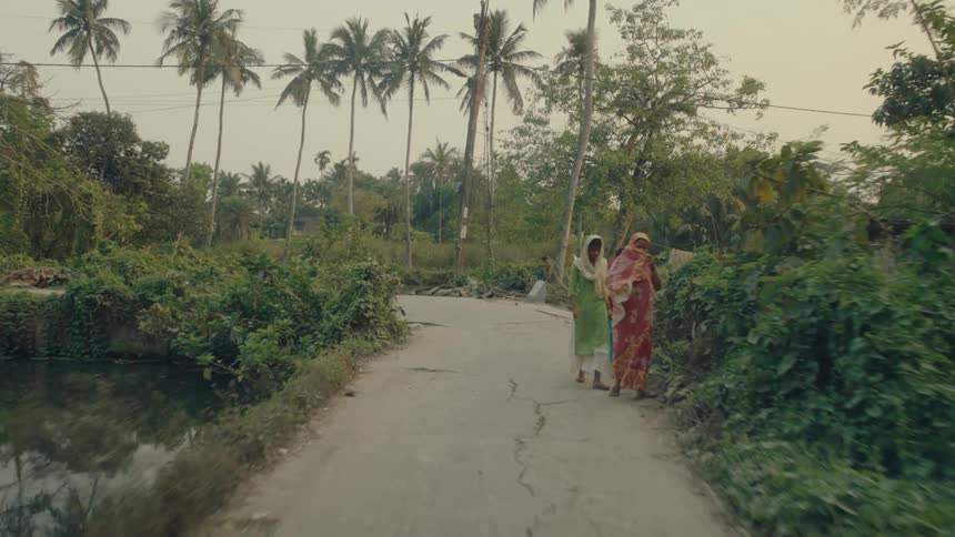 A narrow unkept road by a river in india with three women walking by and covering their faces