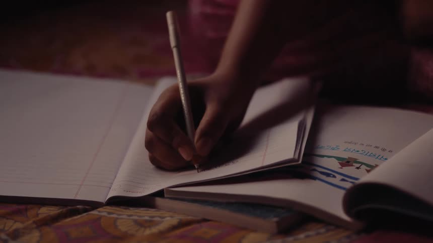 A child writes in a notebook while sitting indoors surrounded by other open books