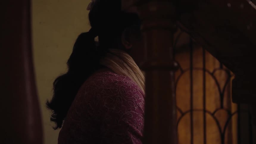 A woman is partially visible in profile sitting or standing indoors near a staircase with her face obscured in shadow