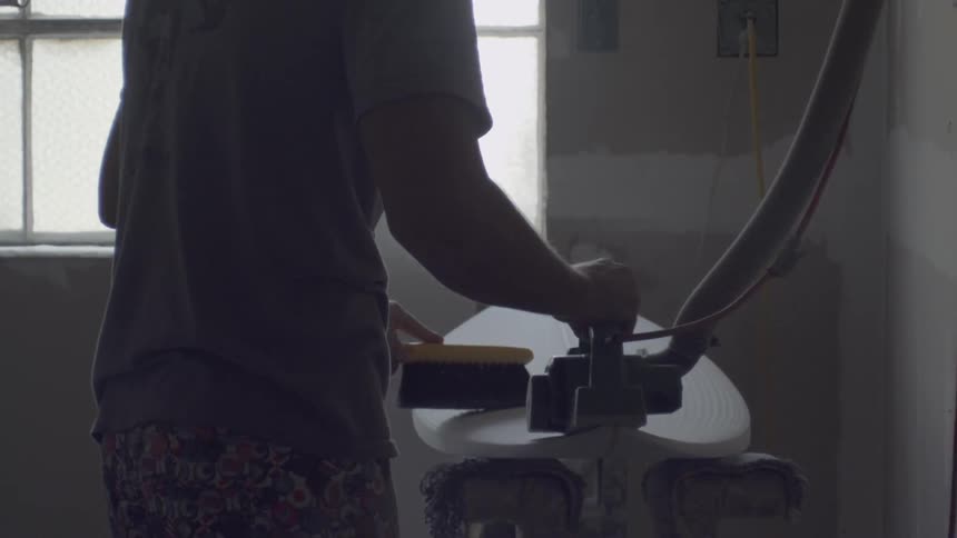 A man uses a tool and brush on an unfinished surfboard