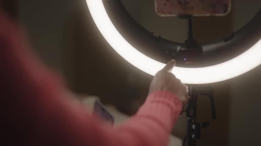 Woman turning on a ring light in her room the ring light is holding a smartphone and a laptop is on the table beside her