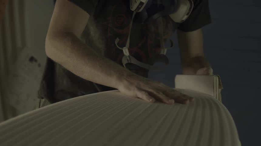 A man is using a sanding block along the edges of a surfboard