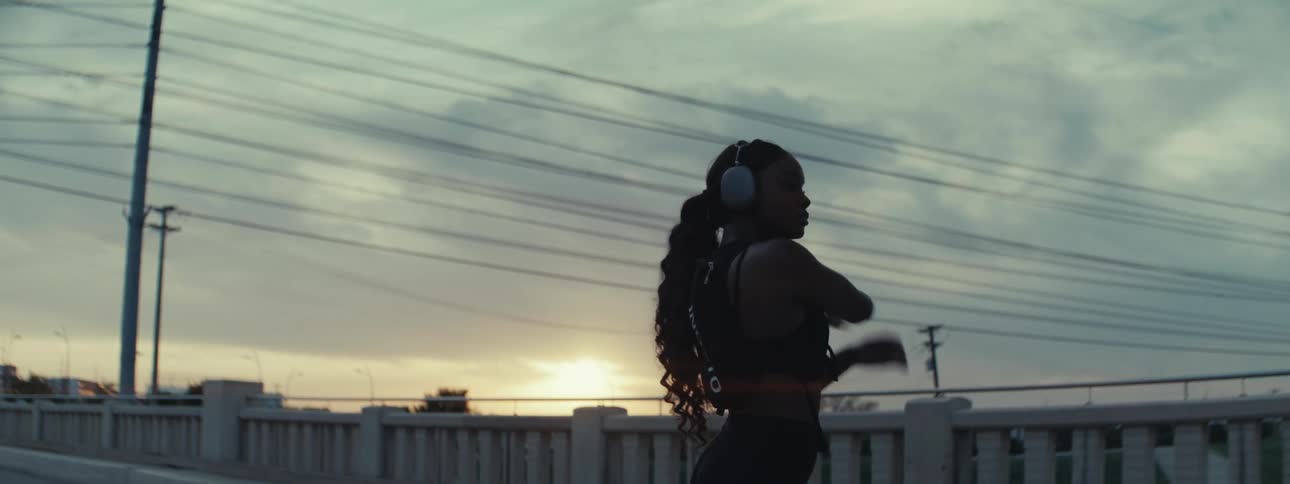 A woman with long hair wearing headphones stretches by the roadside at sundown