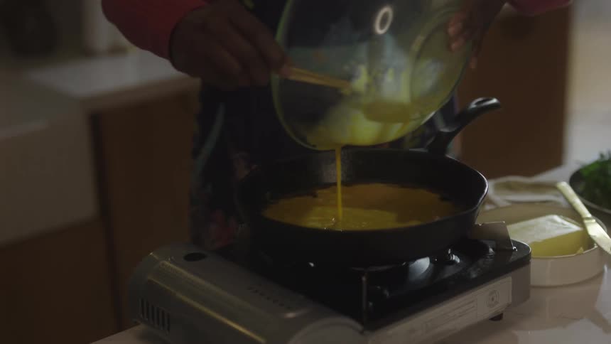 A woman in a floral apron pours beaten eggs into a frying pan set on a portable burner while preparing a meal in her kitchen