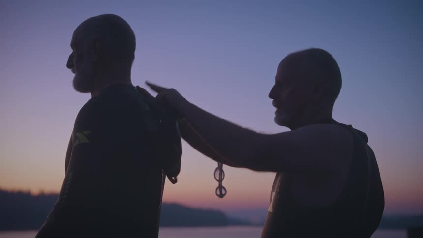 A man wearing a divers suit pats another man on the back during sunset both silhouetted against the warm fading light
