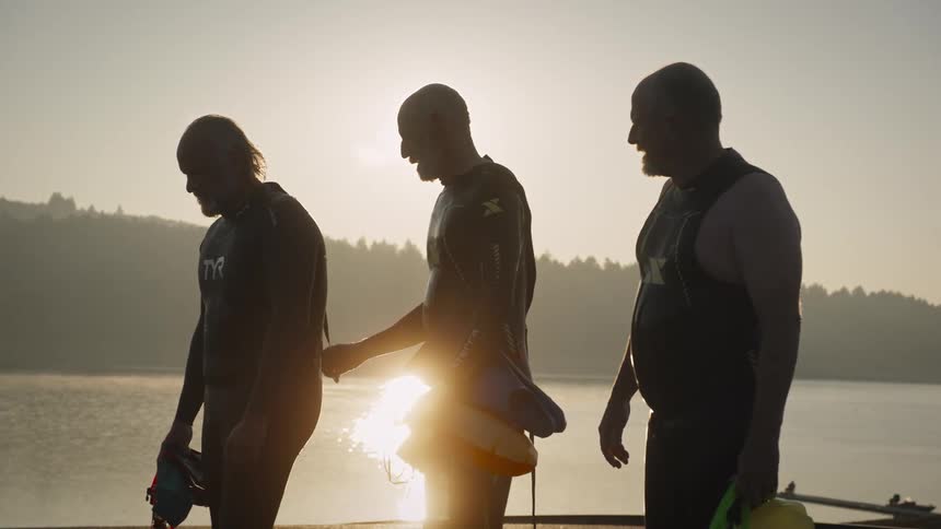 Three adult men in wetsuits walk along a lakeside at sunrise or sunset silhouetted against the shimmering water likely having just completed or preparing for an openwater swim