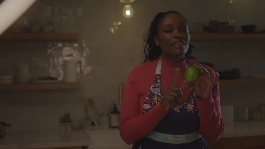 A woman stands in a warmly lit kitchen holding a lime and smiling as she appears to explain or present something on camera