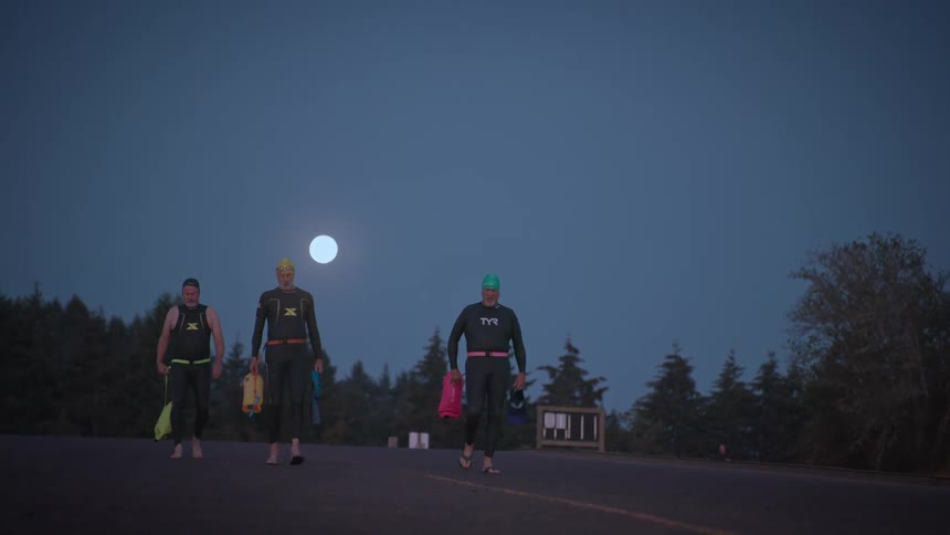 Three people in wetsuits walk barefoot on a road at dusk or dawn likely heading to or from a swim with the moon visible in the background