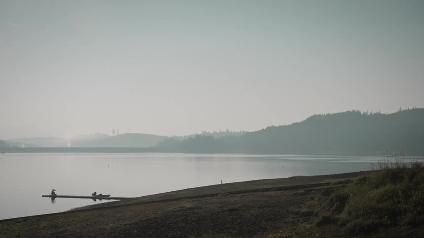 Two people sit peacefully at the end of a dock on a calm lake surrounded by misty hills and still water under a hazy sky