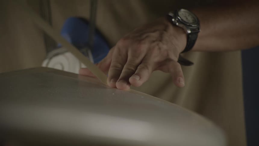 A man is applying a strip of masking tape to a surfboard in a workshop