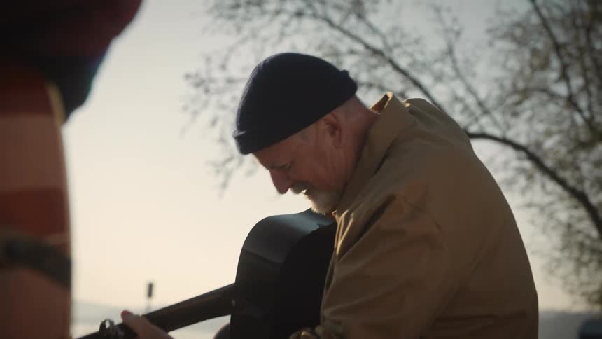 A person wearing a beanie and a brown jacket plays an acoustic guitar outdoors near a lake with trees in the background