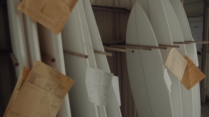 Unfinished surfboards with order tags attached line the walls of a workshop