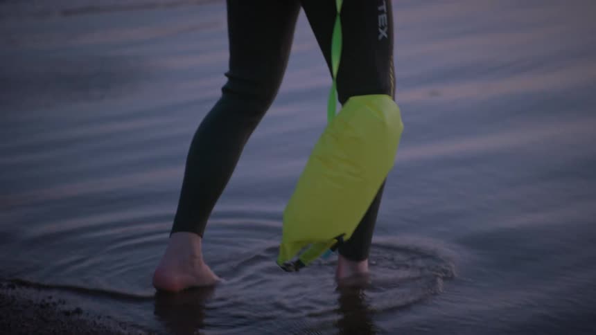An elderly man in a wet suit and a swimming cap with a safety buoy attached to him via a cord steps carefully into the sea near a pontoon