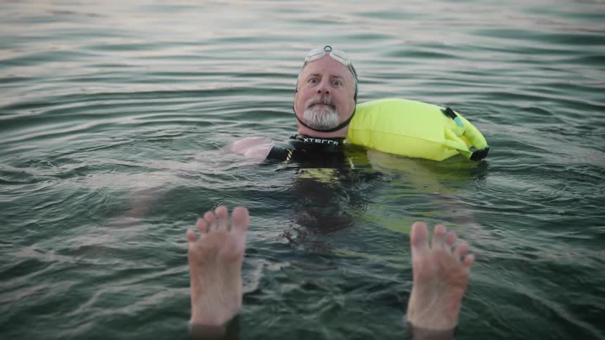 A middleaged man floats in open water with swim goggles and a yellow flotation device as another persons feet in the foreground hint at a shared swim