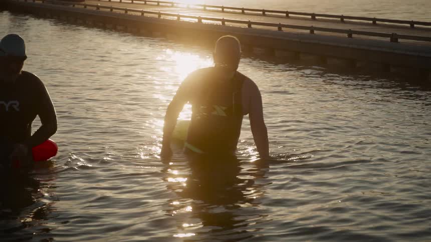 Two people wade through shallow water at sunset with golden light reflecting off the surface as they move near a bridge or pier
