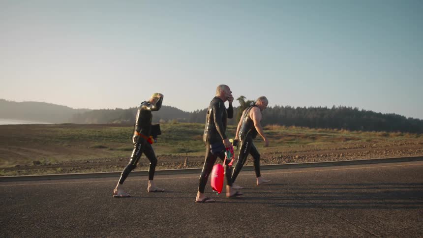 Three people in wetsuits walk barefoot on a road toward a body of water carrying swim gear during early morning light