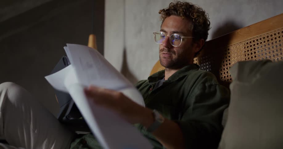 Man sitting on his bed looking over documents while working on his laptop
