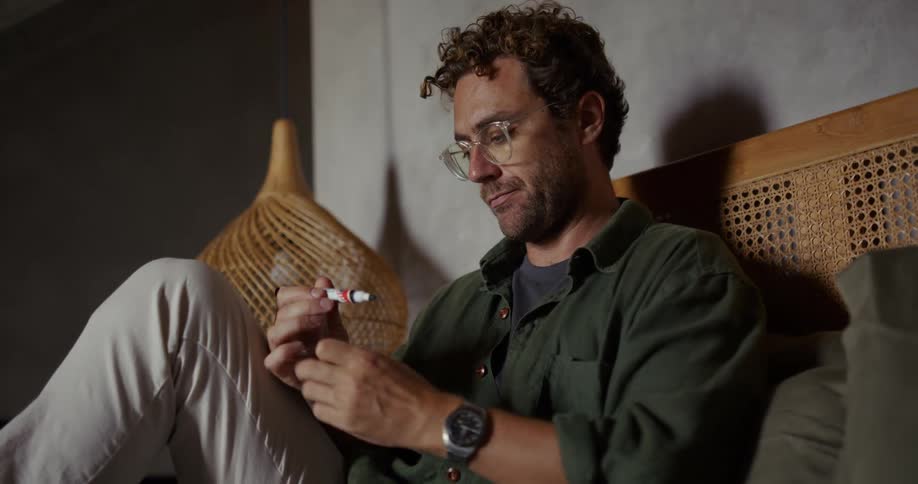 A young man wearing glasses sits on a bed in a bedroom using a marker to label small containers filled with water samples