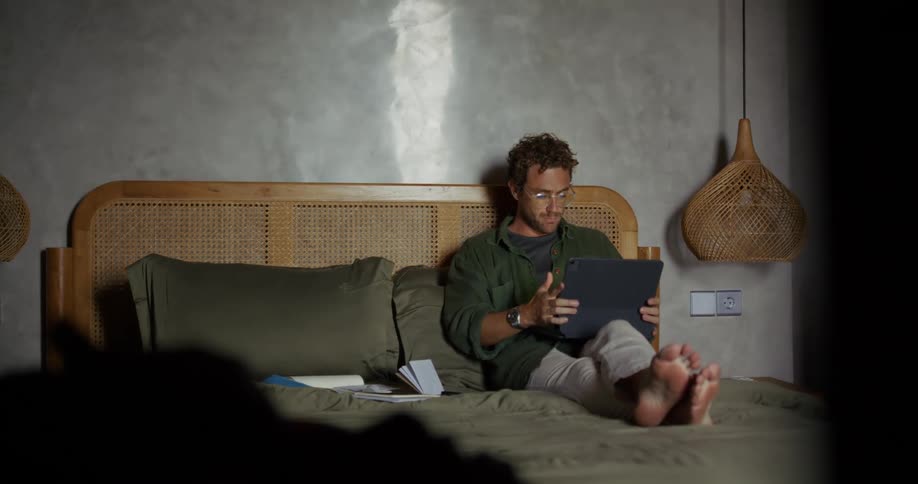 Man with glasses sitting on bed and picking up a tablet