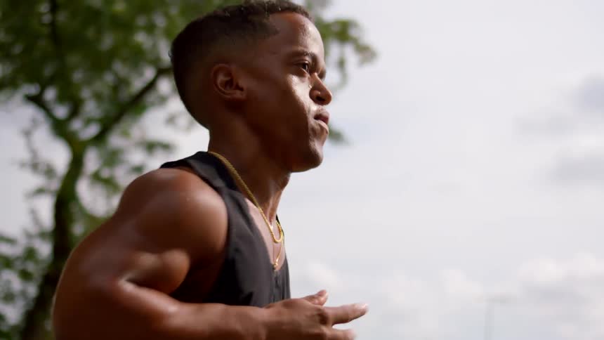 A little person wearing a black sleeveless shirt and a gold necklace jogs outdoors with trees and a cloudy sky in the background