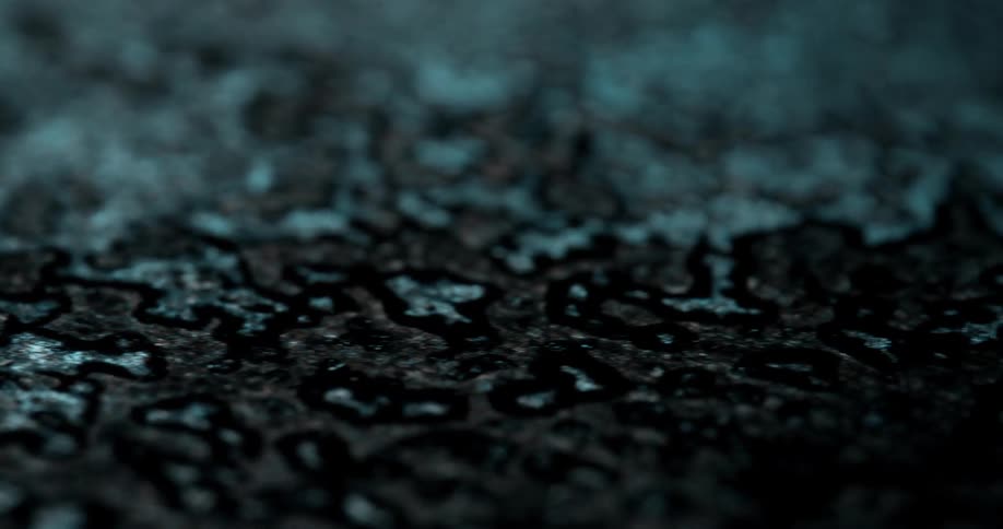 Dark abstract blotches resembling ink or corrosion patterns spread across a textured blue surface in an extreme closeup
