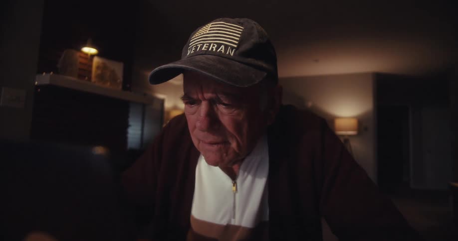 An elderly man in a veteran cap sits at a laptop in a warmly lit home gradually shifting from focused concentration to a gentle emotional smile