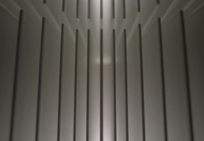Wall with vertical metal panels is seen in perspective with centralized lighting at the top