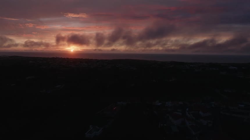 An aerial view shows a vibrant sunset over the ocean with clouds painted in warm colors above a silhouetted coastal landscape dotted with a few lights