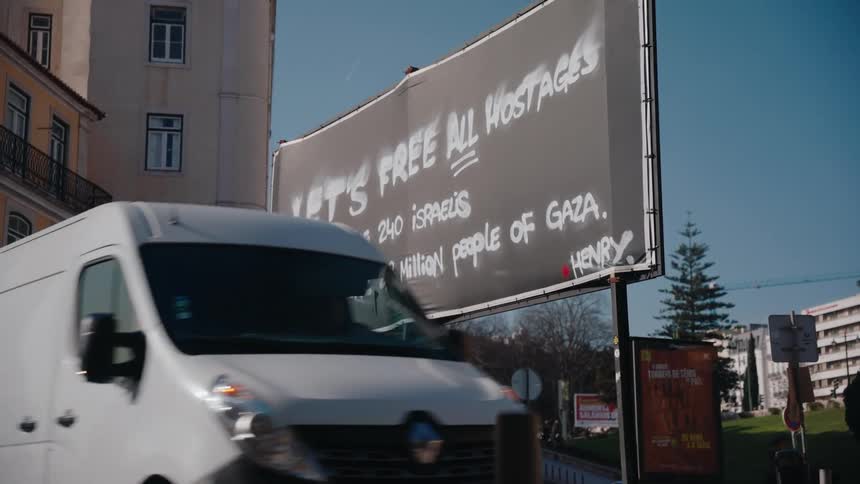 A protest sign on a billboard in a busy city street with a van passing in front and a nearby road filled with cars in transit