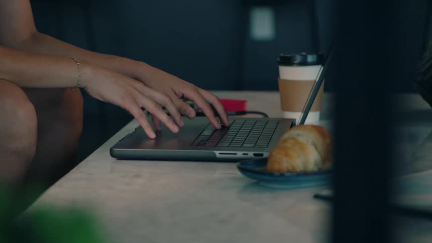 A croissant and a coffee cup sit on a table with a laptop in between them as someone continuously types on the laptop keyboard