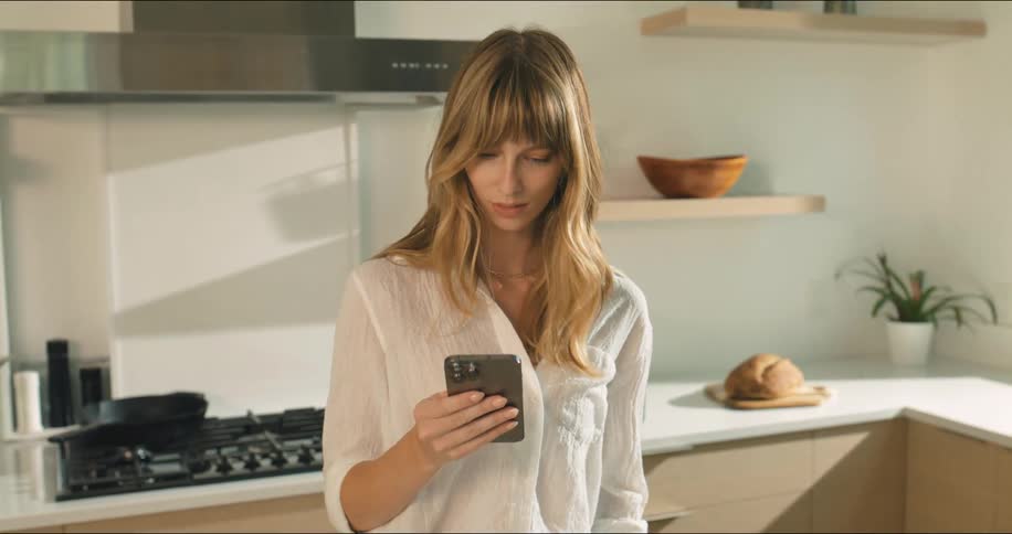 A woman cutting asparagus in the kitchen then pausing to look at her phone and smiling as she sees something on the screen