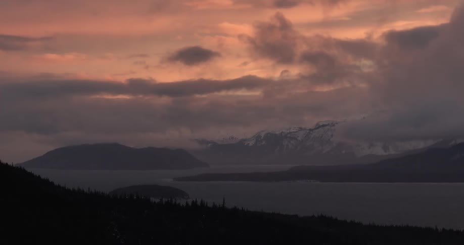 A serene landscape features a body of water with islands and snowcapped mountains in the distance all under a sky filled with soft colorful clouds