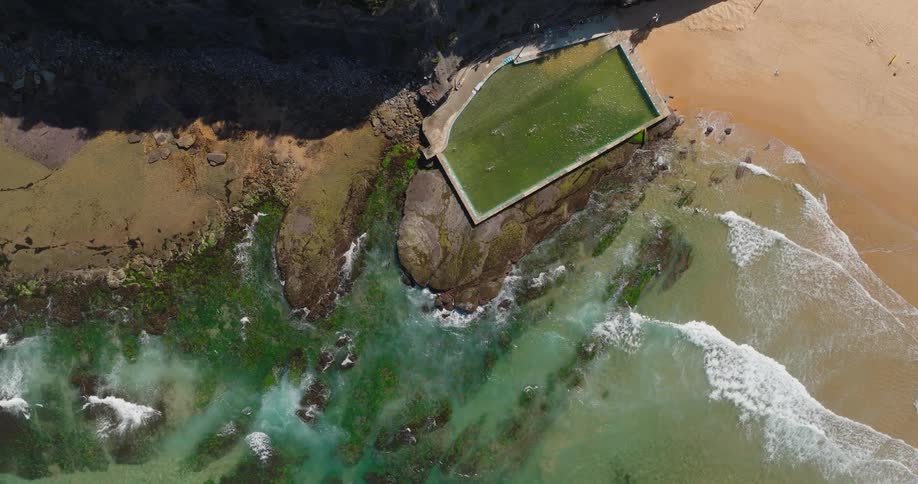 An aerial view shows a rectangular greenfilled swimming pool nestled between rocky outcrops and a sandy beach
