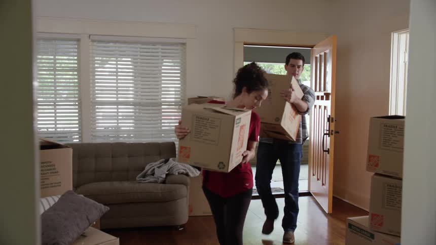 A man and woman carrying boxes enter a house