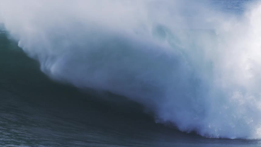 A massive ocean wave curls and breaks in dramatic slow motion sending plumes of white spray and sea foam into the air as the dark water crashes down