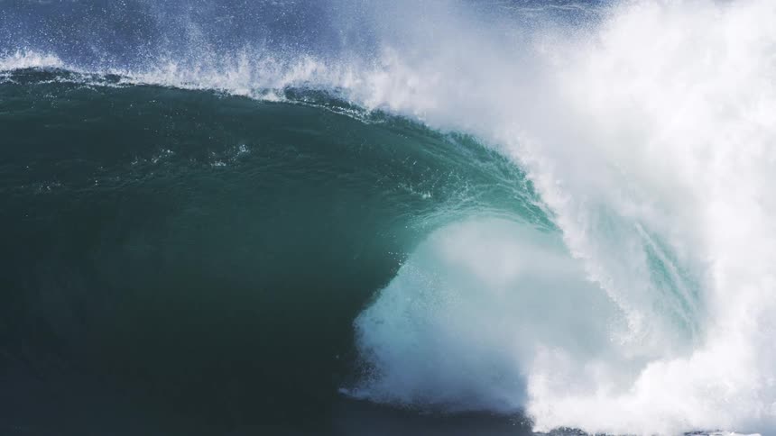 A massive dark blue ocean wave curls and breaks dramatically in slow motion sending plumes of white sea spray high into the air