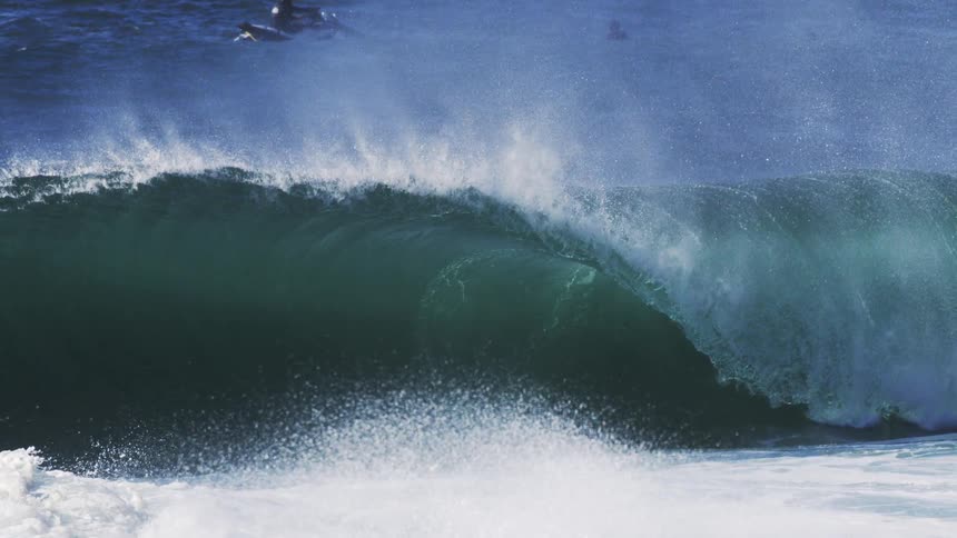 A surfer floats in the ocean as a wave begins to form under the bright midday sun