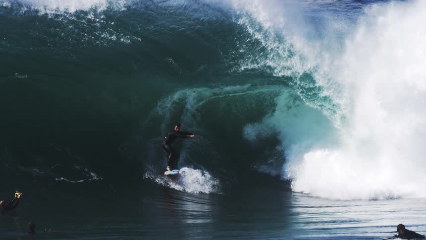 A person surfs a gigantic ocean wave next to two other people who are already bobbing in the water below