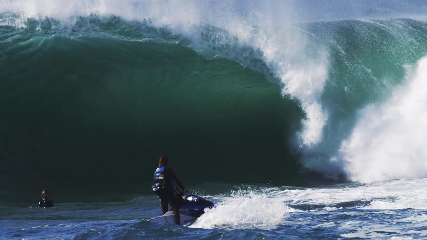 A person on a jet ski navigates near a towering wave while another individual is visible in the water nearby
