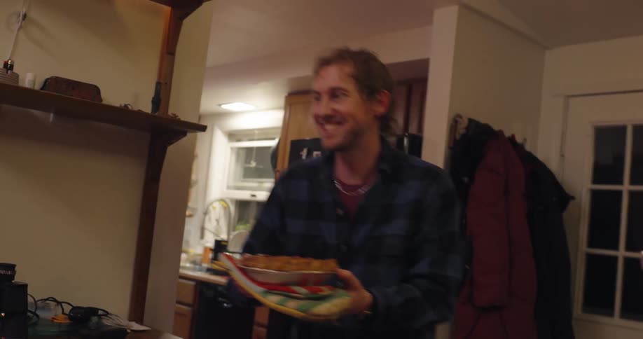 A smiling man carries a pie through a cozy kitchen and into a dinning room presenting it proudly while wearing a plaid shirt in a warmly lit home interior