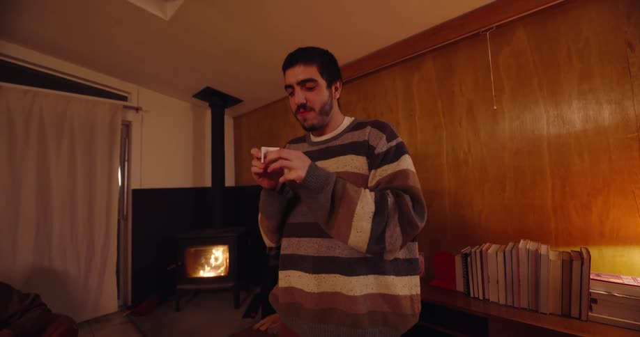 A man wearing a winter sweatshirt stands in front of a group of friends sitting in the living room at night with a lit fireplace behind him as he picks a piece of paper from a bowl on the table and gestures animatedly while demonstrating
