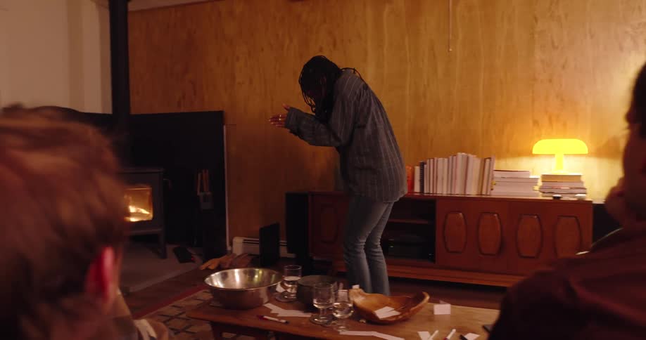 A black woman demonstrates a word in front of a group of friends as they play charades in a living room at night