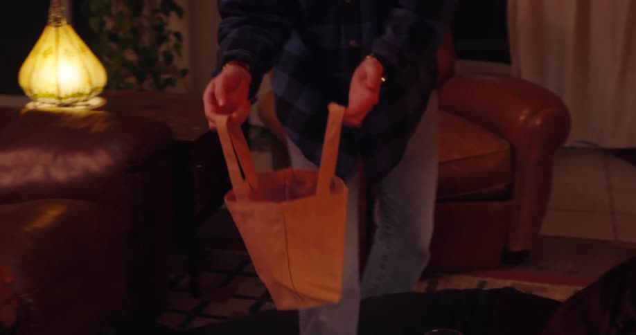 In a warmly lit room with a fireplace a man encourages his friends to place their cellphones into a bag