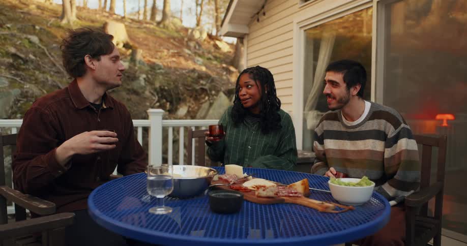 Three friends chat casually over drinks and snacks on a forestside patio