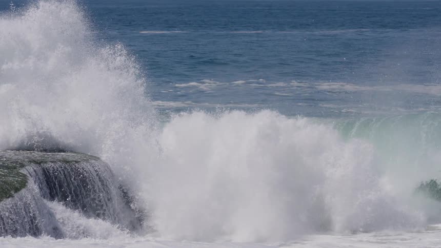 A large ocean wave crashes powerfully against dark rocks sending up significant white spray while water also cascades over a flatter rock in the foreground